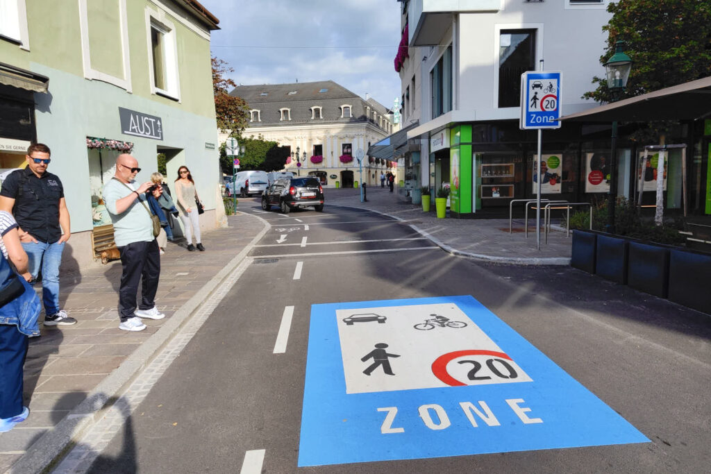 Straße in Tulln Ort mit niedrigen Wohnhäusern, Bodenmarkierung und Schild "Begegnungszone". Radfahren ist hier in beiden Richtungen möglich (durch den Mehrzweckstreifen ersichtlich). Autos dürften hier nur in Blickrichtung in der Begegnungszone unterwegs sein.