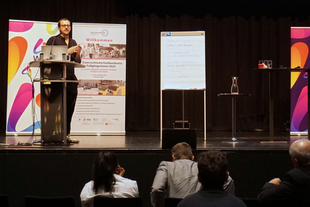 Der Vortragende Jonathan Fetka (Brille, schwarz gekleidet) steht auf der Bühne. Auf der Bühne befinden sich Rollup Banner mit Informationen, sowie eine beschriebene Flip Chart und ein Rednerpult mit Laptop und Mikrofon. Weitere Stehtische mit Getränken stehen auf der Bühne. Eine Leinwand auf die der Vortrag projiziert wird ist angeschnitten zu sehen. Im Vordergrund erkennt man das sitzende Publikum.