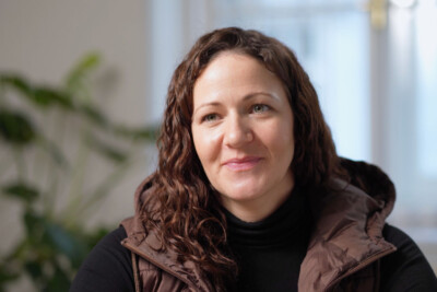 Frau (Portrait) mit braunem Haar, braunem ärmelloser Kapuzenjacke und schwarzem Pullover, in einem Raum. Im Hintergrund Zimmerpflanze und Fenster