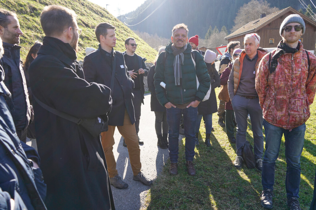 Gegenlichtfoto eine Personengruppe einer Führung auf Weg. Links und Rechts Wiese. Hinten Berge. Winterliche Bekleidung