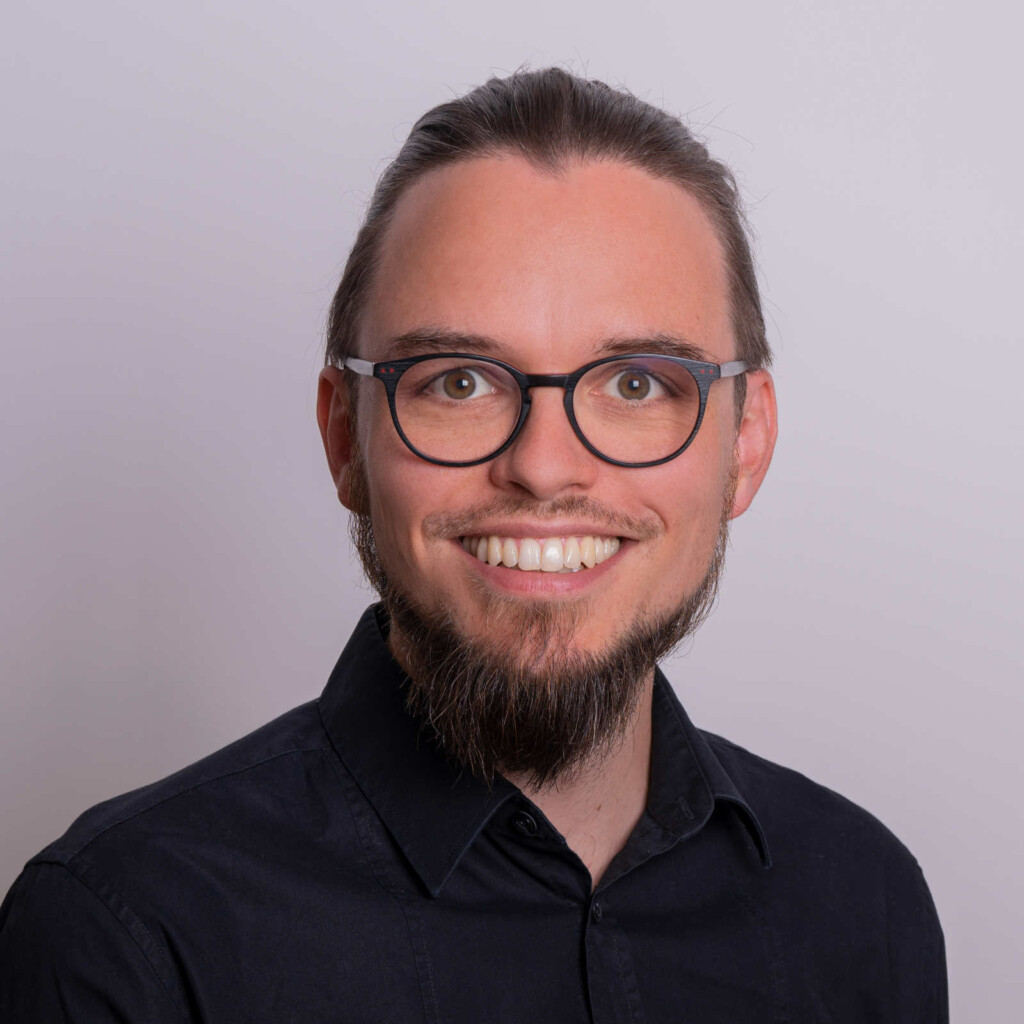 Portrait:Junger Mann mit Brille und streng nach hinten gefassten Haaren. Schwarzes Hemd.