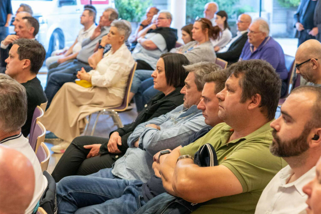 Zahlreiche Bürger:innen sitzen bei einer Veranstaltung auf Stühlen, sommerlich gekleidet