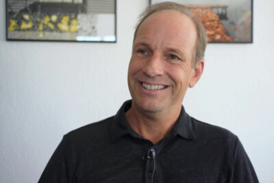 Portrait Dieter Egger. Schwarzes Poloshirt, hohe Stirn, freundliches Lächeln, Hintergrund weiße Wand mit zwei Bildern