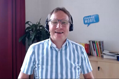 Portrait Ekkehard Allinger Csollich. Mann mit Brille, Kopfhörer, kurzes blau gestreiftes Hemd sitzt im Büro