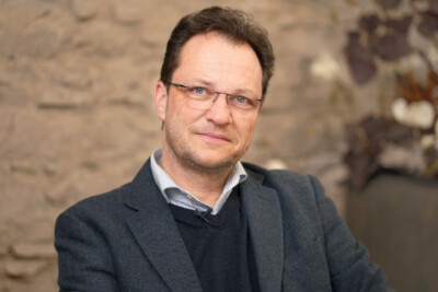 Portrait Jens de Buck. Dunkle Haare, Brille, Sackko, Pullover, offenes Hemd