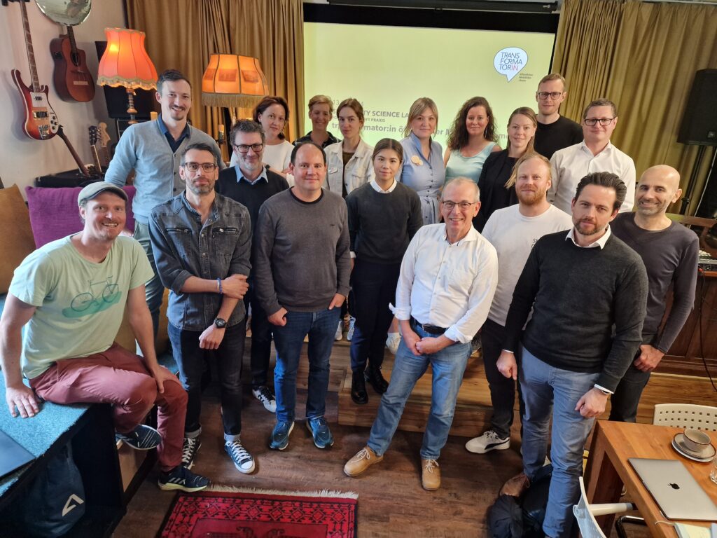 Gruppenbild mit 18 Personen in einem Kulturraum. Vorhang und Gitarren sind im Hintergrund zu sehen.