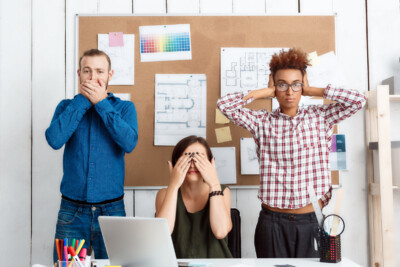 An einem Bürotisch. Drei Personen, einer hält sich den Mund zu, eine die Augen und eine die Ohren. Im Hintergrund an der Wand eine Pinnwand mit Zettel und Plänen.