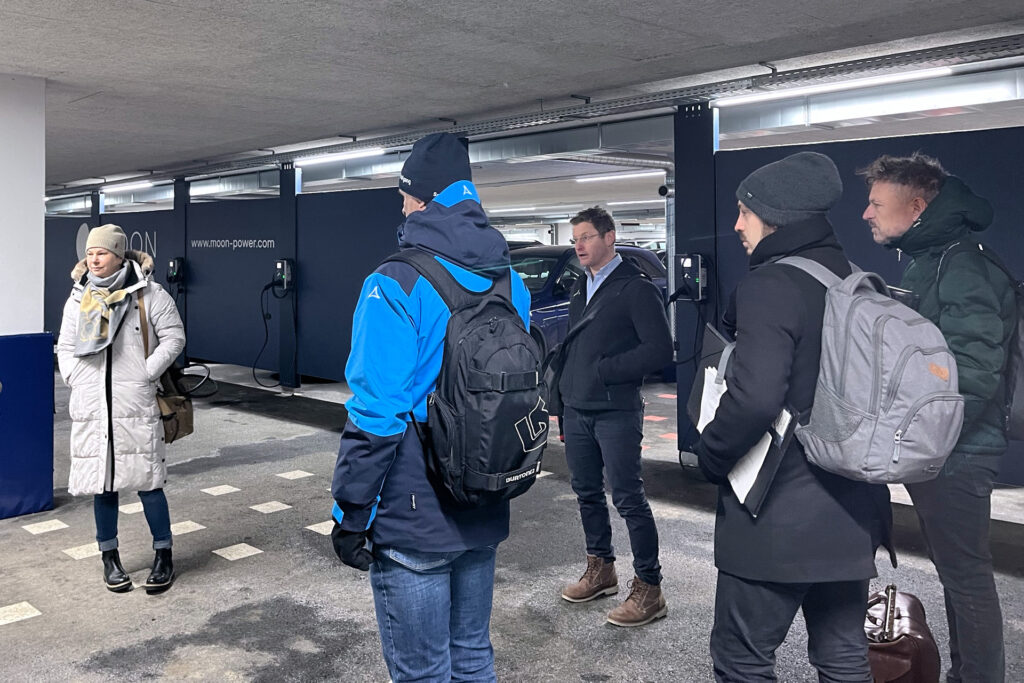 kleine Personengruppe steht in einer Tiefgarage, winterliche Kleidung. An der Wand e-Tankstellensysteme. Ein Mann erklärt etwas.