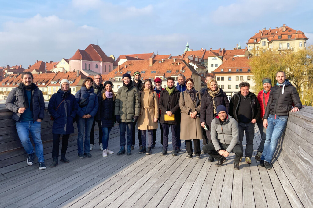 Personengruppe auf Fußbrücke an der Lent. Holzboden. Im Hintergrund Teile der Stadt Maribor.