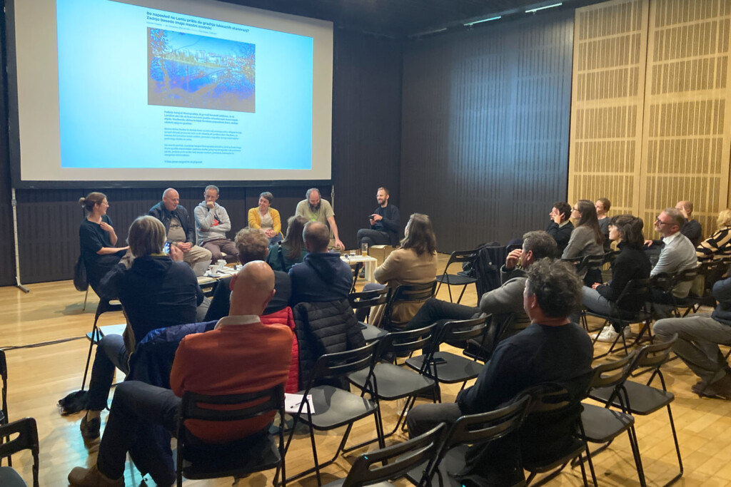 Podiumsdiskussion. 6 Personen am Podium, Publikum. Edler Saal mit dunkler Holzwand und goldfarbener Holzwand. Holzboden