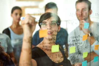 Workshopsituation. Frau notiert auf PostIt an einer Glaswand. 3 Personen stehen interessiert um sie herum.