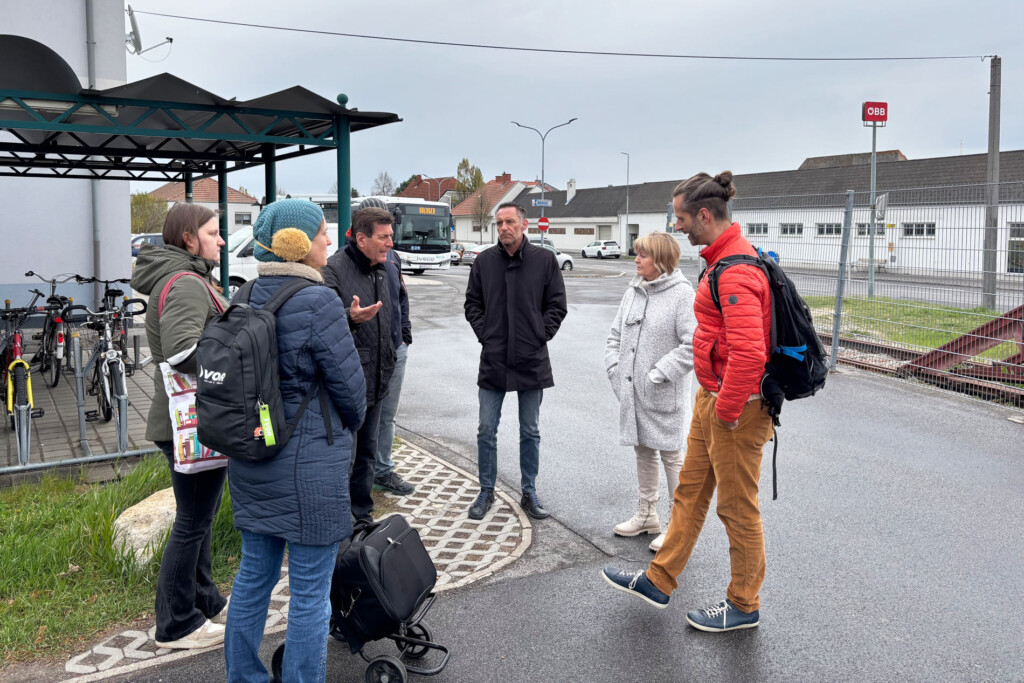 Personengruppe diskutiert vor Bahnhof Deutschkreutz, Im Hintergrund Bahnhofsgebäude und Radabstellanlagen sowie ebenerdige Häuser