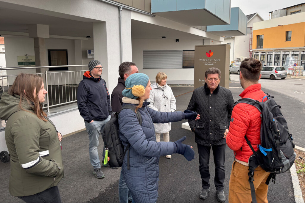 Personengruppe steht vor einem mehrstöckigem Haus ("Wohnen im Alter") eher kreisförmig und tauscht sich aus.