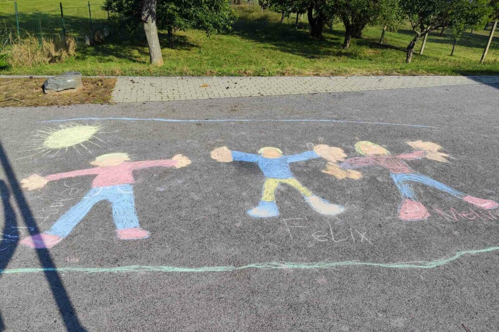 Kinderkreidezeichnung von 3 Personen auf einer Straße. Angrenzend an die Straße eine Streuobstwiese.