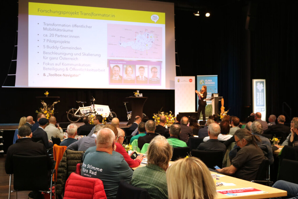 Veranstaltungsraummit Vielzahl an Gästen an Tischen. Eine Frau hält einen Vortrag. Eine Folie "Forschungsprojekt Transformatorin" ist an die Projektswand gebeamt.