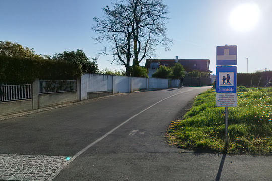 ländliche schmale Straße. Einfamlienhäuser an einer linken Seite. An der anderen Seite ein Gehsteig anfolgend eine Wiese. Verkehrsschilder Sackgasse, darunter Schulstraße