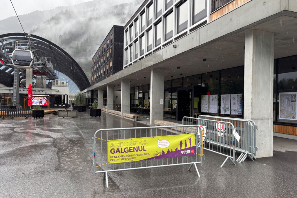 Regnerischer Tag. Talstation, moderner Hotelkomplex. Nebel auf den Bergen im Hintergrund. Im Vordergrund: Ein Transparent: Aktiontag Galgenul. Deine Ideen für den Gestaltung des Ortsteilplanes in auffälliger Farbgebung.