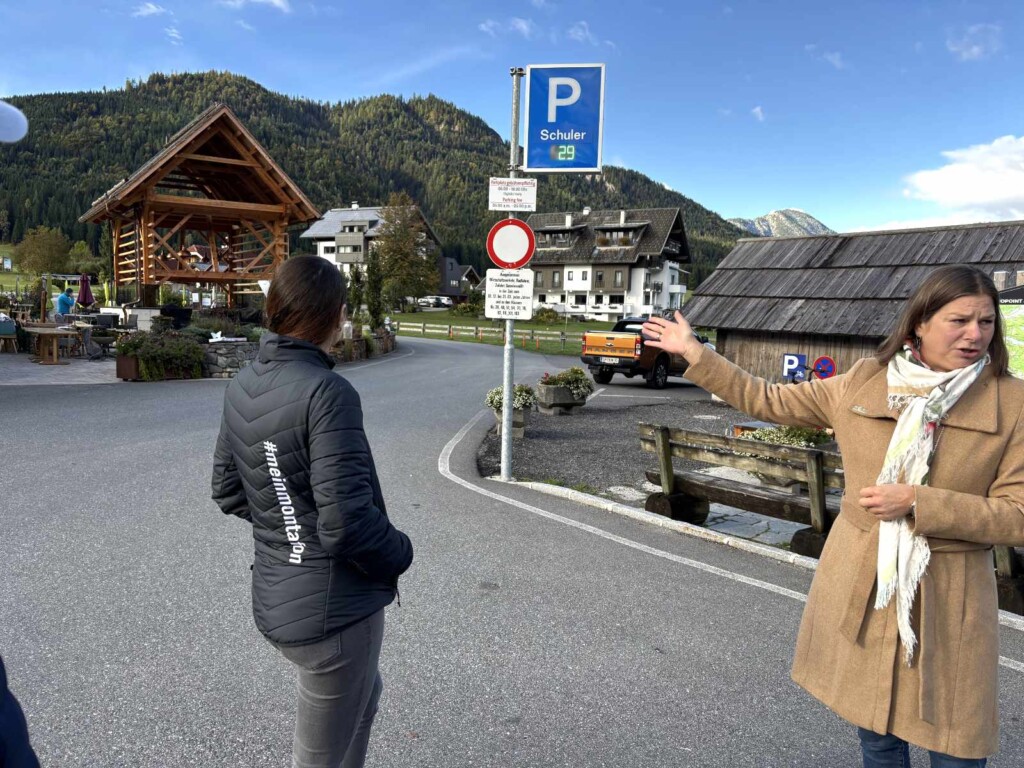 Eine Frau in hellbraunem Mantel steht auf einer Straße (im rechten Bildrand) und deutet auf den Außenbereich eines Restaurant. Eine Frau in schwarzer Jacke steht mit dem Rücken zur Kamera neben ihr und blickt auf eine Straße, die Richtung bewaldete Berge führt. Rechts von der Straße stehen Häuser mit Schieferdächern. Im Mittelpunkt des Bildes steht ein Parkplatzschild. Darauf steht "Schuler".