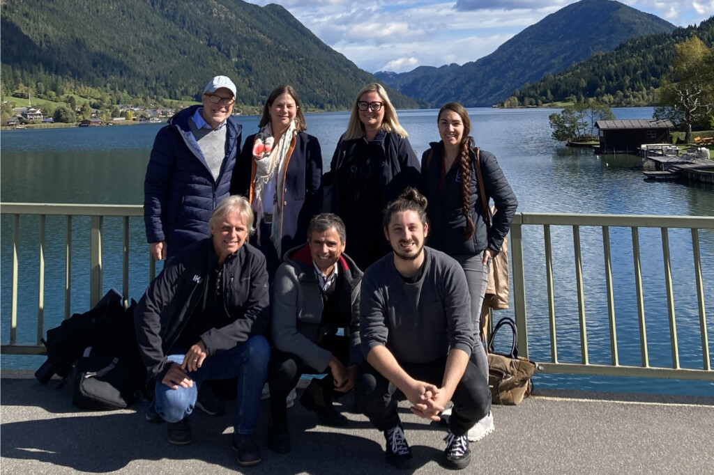Eine Gruppe Menschen steht auf einer Brücke und lächelt in die Kamera. Hinter ihnen ist ein See, eingerahmt von Bergen, zu sehen.