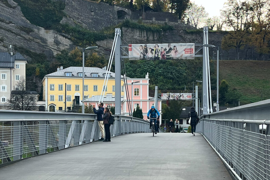 Rad- und Fußgängerbrücke über die Salzach mit Passanten und einem Radfahrer im Hintergrund traditionelle Salzburger Bauten und der Mönchsberg