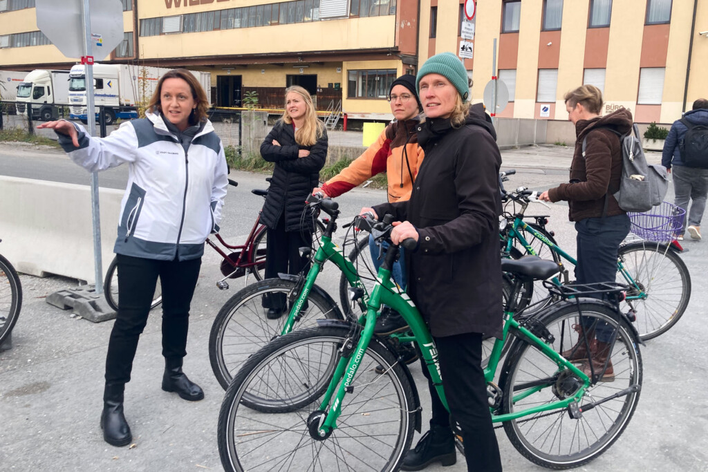 weibliche Personengruppe mit Fahrrädern blickt in eine Richtung, in die die Vortragende zeigt. Im Hintergrund einige jahrzehnte alte gewerbliche Industriebauten