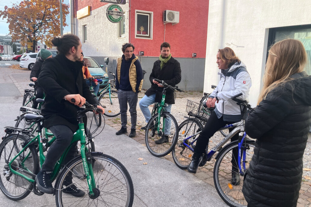 Gruppe von radfahrenden Personen steht im Halbkreis, eine Person trägt vor. Im Hintergrund kleines Haus mit roter Fassade mit einem Gastronomiebetrieb