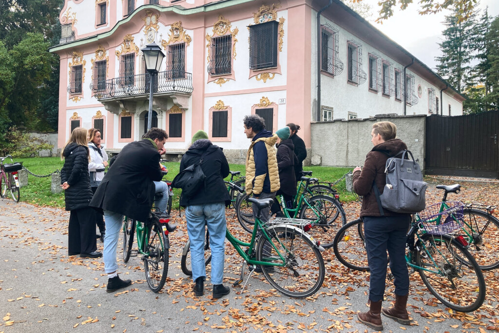 Radfahrgruppe steht vor einem barocken kleinem Schloßartigen Gebäude, dieses ist von Grün und Bäumen umgeben.