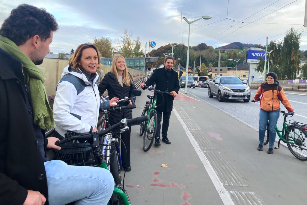 Radfahrgruppe steht an einer stark befahrenen Straße auf einem Gehsteig auf dem auch ein Radweg markiert ist.