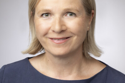 Portraitfoto: Eine Frau mit hellblonden, halblangen Haaren und dunkelblauem U-Boot-Shirt, grün-braunen Augen, lächelnd.