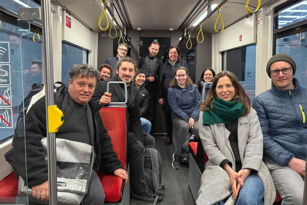 Viele Menschen schauen in einer Straßenbahn in die Kamera. Die meisten sitzen.