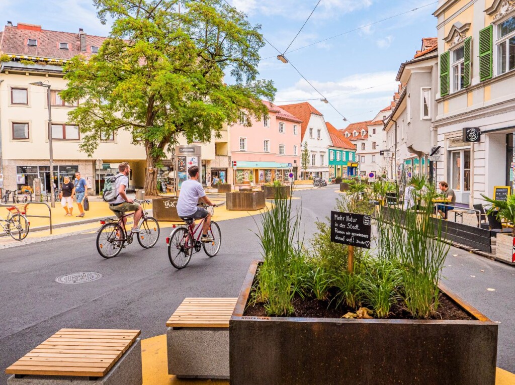 Zwei Radfahrende auf einem Platz, gesäumt von ein und zweistöckigen Häusern. Am Boden strukturieren größere und kleinere, goldgelbe Kreisflächen den Verkehrsraum. Auf den goldgelben Flächen stehen große Pflanztöpfe aus Edelrost mit Bäumen und anderen Pflanzen. Sitzgelegenheiten auf den gelben Flächen laden zum Verweilen ein. Ein großer Baum unterstützt die angenehme Athmosphäre des öffentlichen Raumes, auf dem in der aktuellen Situation auch keine Autos sichtbar sind.