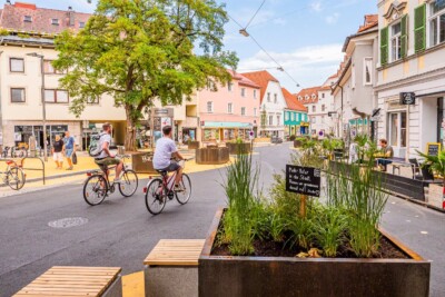 Zwei Radfahrende auf einem Platz, gesäumt von ein und zweistöckigen Häusern. Am Boden strukturieren größere und kleinere, goldgelbe Kreisflächen den Verkehrsraum. Auf den goldgelben Flächen stehen große Pflanztöpfe aus Edelrost mit Bäumen und anderen Pflanzen. Sitzgelegenheiten auf den gelben Flächen laden zum Verweilen ein. Ein großer Baum unterstützt die angenehme Athmosphäre des öffentlichen Raumes, auf dem in der aktuellen Situation auch keine Autos sichtbar sind.