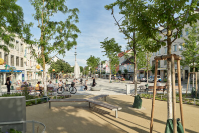 Eine Sitzbank, umgeben von Bäumen auf einem weitläufigen Platz in einer Stadt. Weiter hinten sitzen Menschen und spielen Kinder vor eingeklappten Sonnenschirmen. Es scheint die Sonne.