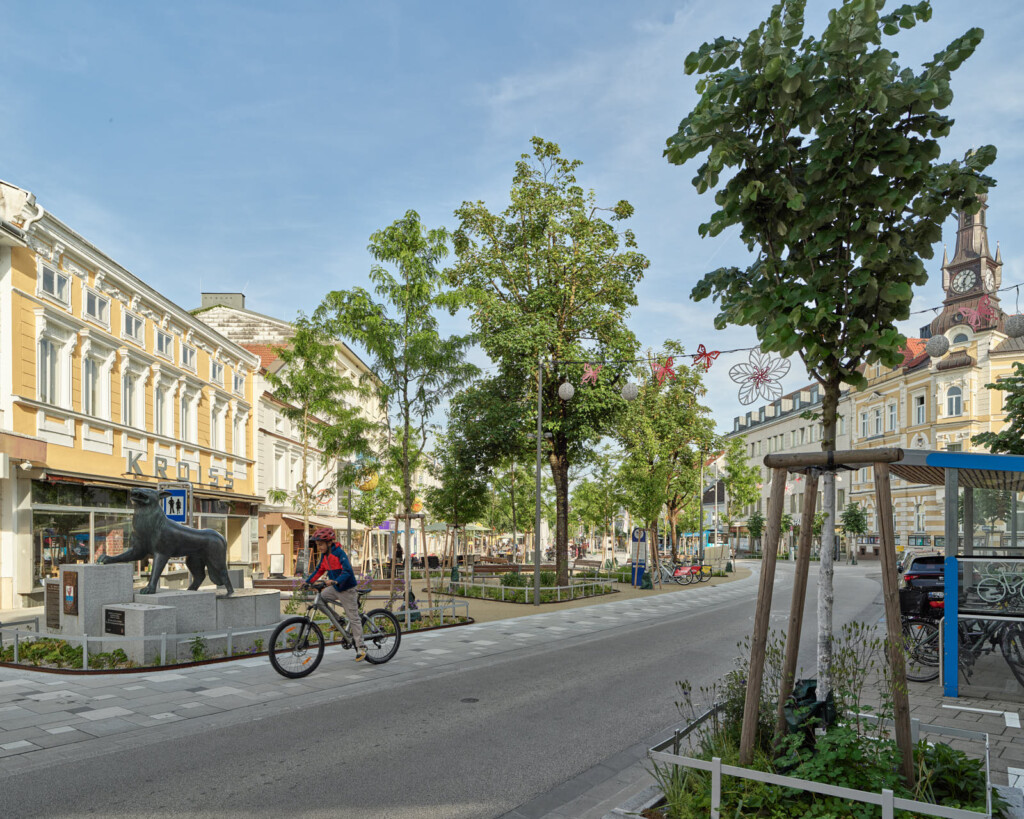 Junge am Fahrrad fährt auf der Straße am Platz einer Kleinstadt mit zahlreichen Bäumen