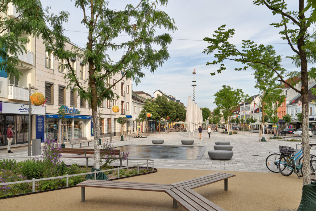 Im Vordergrund eine sternförmige Sitzgelegenheit aus Holz. Blick auf den Platz mit Wasserpsiel und zahlreichen Bäumen. Boden mit Betonsteinen gestaltet