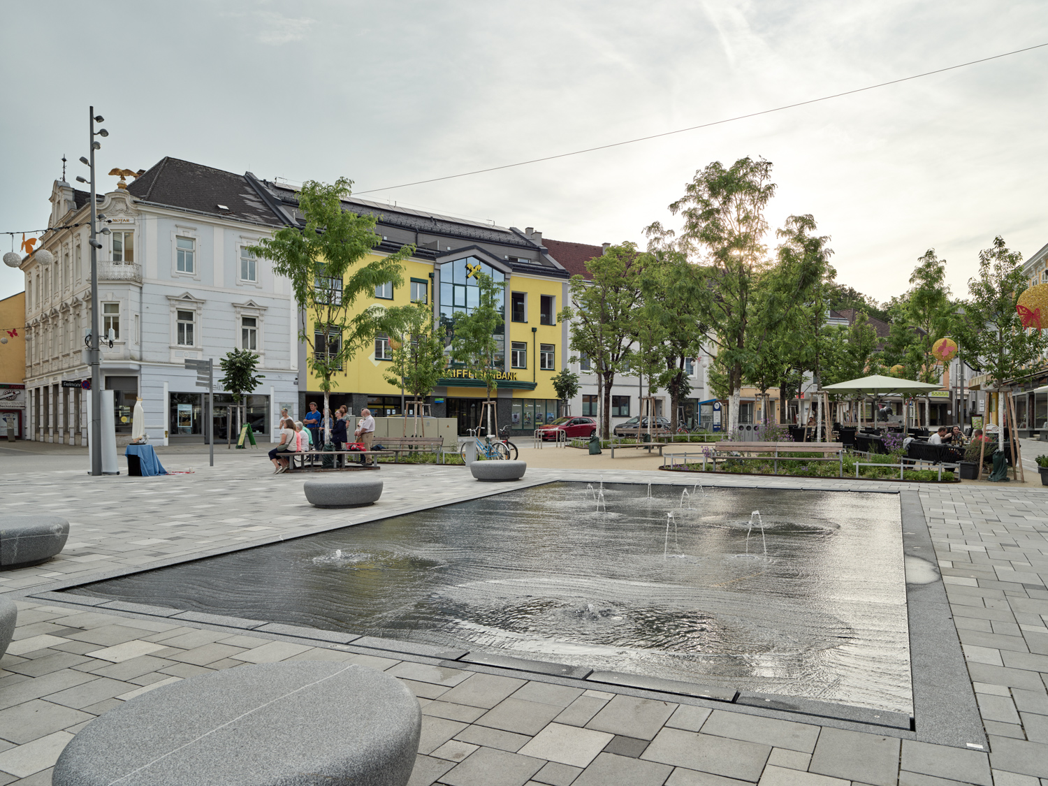 Zu sehen ist ein bodenebener Wasserbrunnen mit kleinen Wasserfontainen auf einem gepflasterten Platz. Im Hintergrund stehen Bäume, dazwischen befinden sich Sitzgelegenheiten. Der Platz wird von zweistöckigen Häusern gerahmt. der Himmel ist wolkenlos. Um einen Baum links neben dem Brunnen sitzen ein paar Menschen.