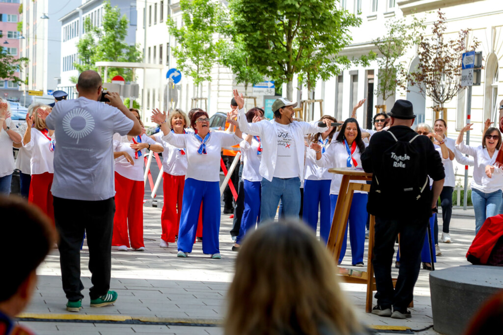 Tanzende Gruppe Erwachsener, meist Damen mit weißen Oberteilen und kräftig Blau bzw. kräftig roten weiten Hosen. Ein junger Mann mit feinem Vollbart ist der Vortänzer und Animator der Gruppe. Im Hintergrund zahlreiche jüngere Bäume und Hausfassaden mehrstöckiger, urbaner Wohnbauten