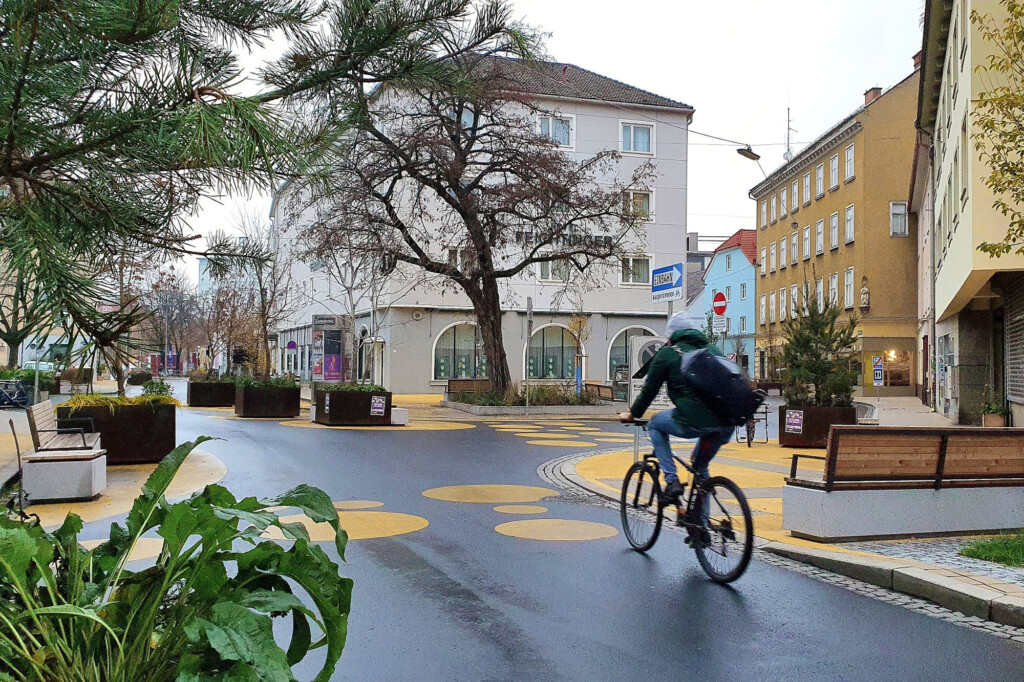 Eine Straßenkreuzung mit orangnen Kreisen in einer Altstadt ist nass vom Regen. Auf ihr fährt im Vordergrund des Bildes eine Fahrradfahrerin. Recht und links von der Straße stehen Pflanzenkübel und einzelne Bänke. Im Hintergrund stehen Bäume und dreistöckige Häuser.
