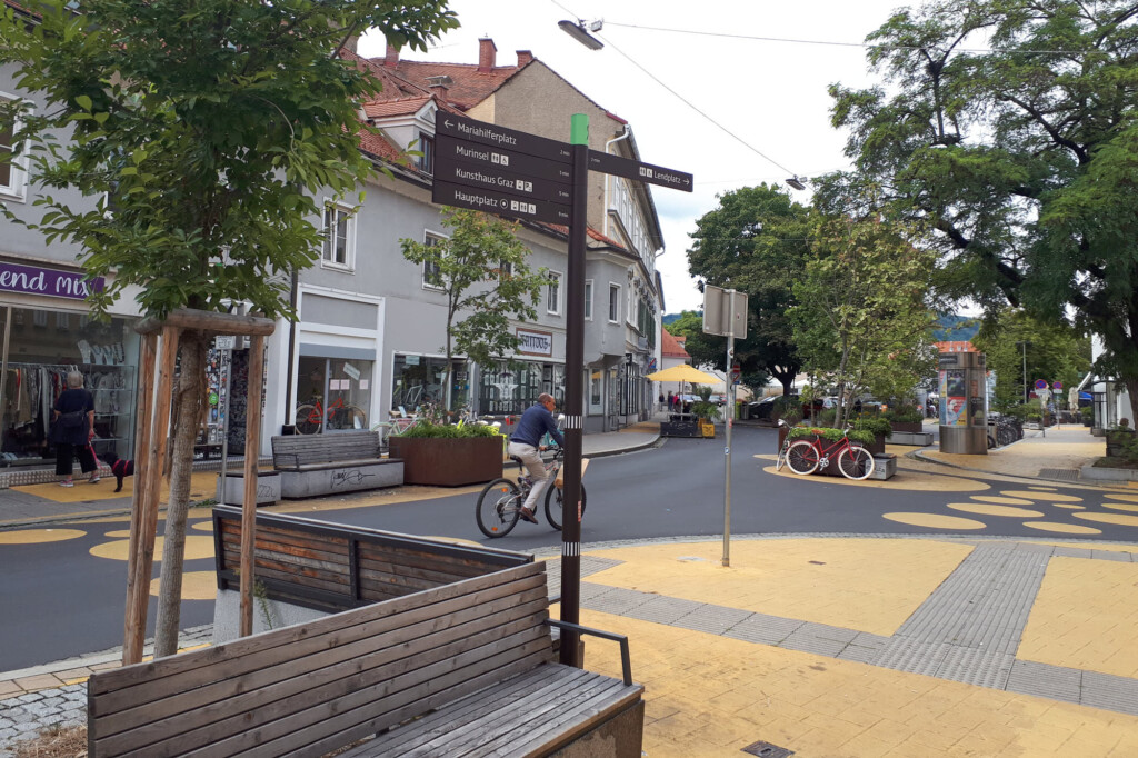 Eine Fahrradstraßenkreuzung mit orangefarbenen Flächen und Kreisen und einem Radfahrer verläuft durch die Altstadt von Graz. Sie ist begrünt und bietet Sitzgelegenheiten. Links von ihr stehen einladende Häuser mit Geschäften im Erdgeschoss. Mittig steht ein Wegweiser, der wichtige Orte anzeigt.