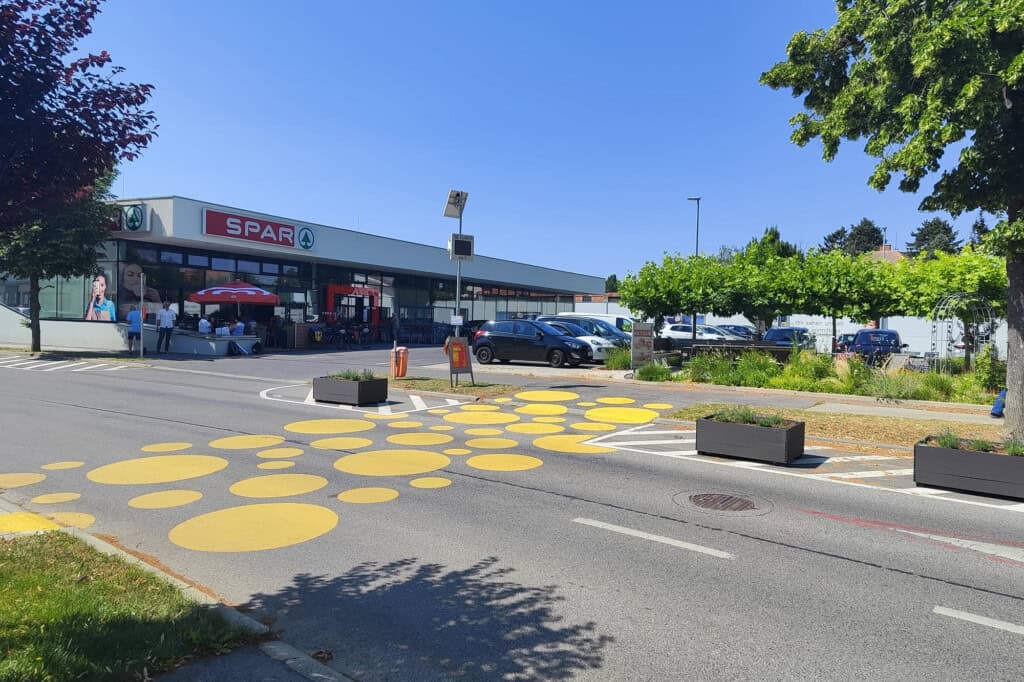 Eine zweispurige Straße, über die eine Querungshilfe aus unterschiedlich großen gelben Kreisen führt. Auf der vom Betrachter abgewandten Seite der Fahrbahn stehen drei Pflanztröge. Im Hintergrund ein großer einstöckiger Supermarkt mit der Aufschrift "Spar"