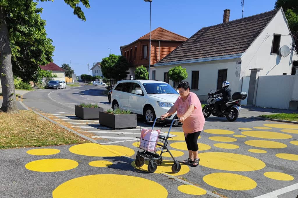 Eine ältere Frau mit Rollator überquert die Straße auf einer Querungshilfe innerorts. Gelbe Kreisflächen unterschiedlicher Größe verbinden gegenüberliegende Gehsteige. Pflanztröge stehen am Fahrbahnrand und flankieren die Querungshilfe. Im Hintergrund einstöckige Häuser. Bäume säumen die Straße. Ein Auto hält, damit die Fußgängerin ungehindert queren kann.