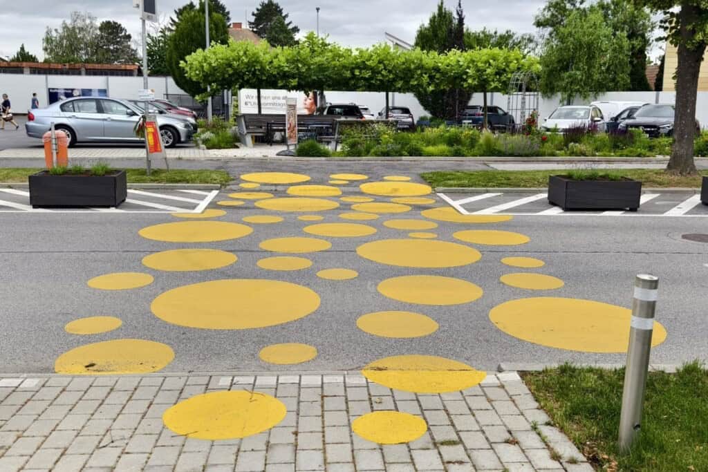 Unterschiedlich große gelbe Kreise führen von einem gepflasterten Gehweg über die Fahrbahn auf die andere Seite. Rechts und links der Querungshilfe stehen zwei Pflanztröge. Im Hintergrund ist ein begrünter Parkplatz zu sehen.