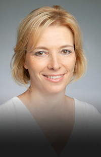 Portraitfoto Barbara Urban. Lächelnd. Brünette, mittellange Haare, weißes Shirt mit V-Ausschnitt.