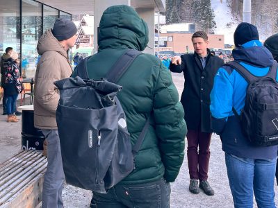 kleine Personengruppe steht im Kreis, winterliche Kleidung, Skitourismusort, Berge und Häuser im Hintergrund schneebedeckt. Ein Mann, Bürgermeister von St.Gallenkirch erklärt einen Sachverhalt.