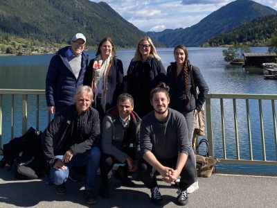 Eine Gruppe Menschen steht auf einer Brücke und lächelt in die Kamera. Hinter ihnen ist ein See, eingerahmt von Bergen, zu sehen.