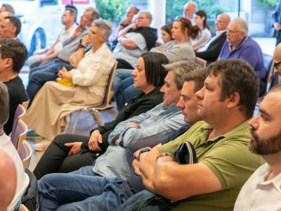 Zahlreiche Bürger:innen sitzen bei einer Veranstaltung auf Stühlen, sommerlich gekleidet