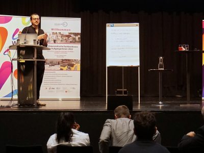 Der Vortragende Jonathan Fetka (Brille, schwarz gekleidet) steht auf der Bühne. Auf der Bühne befinden sich Rollup Banner mit Informationen, sowie eine beschriebene Flip Chart und ein Rednerpult mit Laptop und Mikrofon. Weitere Stehtische mit Getränken stehen auf der Bühne. Eine Leinwand auf die der Vortrag projiziert wird ist angeschnitten zu sehen. Im Vordergrund erkennt man das sitzende Publikum.