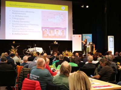 Veranstaltungsraummit Vielzahl an Gästen an Tischen. Eine Frau hält einen Vortrag. Eine Folie "Forschungsprojekt Transformatorin" ist an die Projektswand gebeamt.