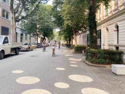 Straße mit Baumallee in Altstadt. Große runde Kreisflächen in Goldgelb sind auf die Straße gemalt. 3 Radfahrende sind unterwegs. Eine Frau sitzt auf einer Bank. Nur ein paar parkende Autos.