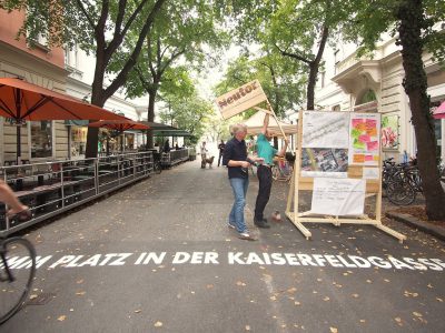 Zum Anlass der Eröffnung der Kaiserfeldgasse wird eine Flagge aus Holz an dem Infostand von 2 Männern befestigt.